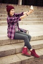 Brunette woman in hipster outfit sitting on steps and taking selfie on retro camera on the street. Toned image Royalty Free Stock Photo