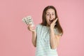 A brunette woman holds a bundle of dollar bills in her hand and covers her mouth with her hand in surprise. Pink background Royalty Free Stock Photo