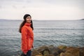 Brunette woman with long hair in a hat and an orange jacket, posing on the beach. In the background the sea and the horizon line. Royalty Free Stock Photo