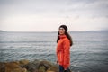 Brunette woman with long hair in a hat and an orange jacket, posing on the beach. In the background the sea and the horizon line. Royalty Free Stock Photo