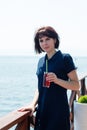 A brunette woman on the pier with glass cherry juice looks away waiting