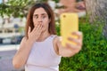 Brunette woman taking a selfie photo with smartphone covering mouth with hand, shocked and afraid for mistake