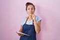 Brunette woman wearing professional waitress apron holding clipboard serious face thinking about question with hand on chin,
