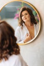 a brunette woman in a white shirt looks at her reflection in a round mirror.