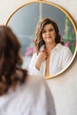 a brunette woman in a white shirt looks at her reflection in a round mirror.