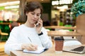 brunette works in a cafe at a laptop talking on the phone writes something in a notebook Royalty Free Stock Photo