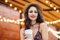 Brunette young lady in summer straw hat and dress, having make-up, holding paper cup with hot drink, having rest at outdoor cafete Royalty Free Stock Photo