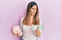 Brunette young woman holding one dollar banknote and piggy bank clueless and confused expression Royalty Free Stock Photo