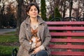 Brunette girl holds small toy Terrier dog in her arms while sitting on red bench in autumn Park Royalty Free Stock Photo