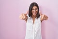 Brunette young woman standing over pink background wearing glasses approving doing positive gesture with hand, thumbs up smiling Royalty Free Stock Photo