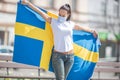 Brunetter standing outdoors looks at the Swedish flag she holds, with a face mask on Royalty Free Stock Photo