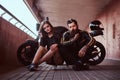 Brutal bearded biker in black leather jacket and sensual brunette girl sitting together on a skateboard near custom-made Royalty Free Stock Photo