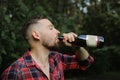 Brutal bearded hipster man drinking wine from the bottle standing outdoors on green trees background. Unhealthy