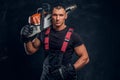 Brutal logger posing with a chainsaw on his shoulder and looking at a camera with a confident look