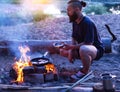 A Brutal Man With A Beard Cooks Food In A Frying Pan On A Fire In Nature. Cooking Bacon On The River Bank. Camping.