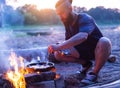 A Brutal Man With A Beard Cooks Food In A Frying Pan On A Fire In Nature. Cooking Bacon On The River Bank. Camping.