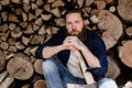 Brutal man with a beard dressed in casual clothes with tattoos on his hands holds the ax sitting near the stacked wood Royalty Free Stock Photo