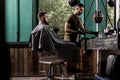 Brutal man with beard sits in a chire at a barber shop. Handsome barber takes the hairclipper