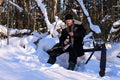 brutal man with beard is sitting on log in winter forest, holding thermos and cup of tea in his hands.