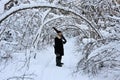 brutal man with beard and in warm sheepskin coat stands among snow-covered forest. Logger is holding an axe and a hand saw.