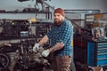 A brutal tattooed bearded mechanic specialist repairs the car engine which is raised on the hydraulic lift in the garage