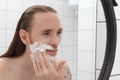 Brutal tattooed man with long hair with shaving foam on his face smiling in front of bathroom mirror Royalty Free Stock Photo