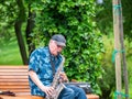 Senior street artist musician sitting on a bench in the park and playing at saxophone Royalty Free Stock Photo