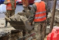 A bucket of a heavy construction excavator surrounded by road workers digs a trench when repairing a city sewer on the roadway