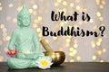 Buddha statue with candle, lotus flowers and singing bowl on table against blurred lights and text What Is Buddhism