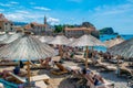 Resting people on sun loungers under thatched umbrellas on Brijeg od Budva beach on background of old city walls fortress. Royalty Free Stock Photo