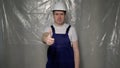 Builder in blue overalls puts on white hard hat and shows thumbs up. Royalty Free Stock Photo