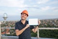 Builder in a protective helmet with a tablet for writing in his hand is standing on the roof of a building overlooking the city Royalty Free Stock Photo