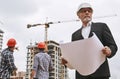 Building inspector. Portrait of experienced elderly engineer in formal wear and white helmet holding a project plan and