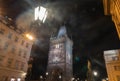 Buildings and statues surround Powder Gates Tower in dark of night with street lanterns illuminating a mistiness and lens flares