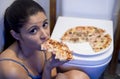 Bulimic woman feeling sick guilty sitting at the floor of the toilet leaning on WC eating pizza