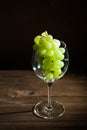 A bunch of green grapes and a wine glass on a dark background in the conditions of artificial hard lighting close up. In a glass o