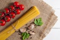 Bunch of raw spaghetti, cherry tomatoes, peppers, garlic cloves and parsley leaves on burlap on a white wooden background.Top view