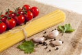 Bunch of raw spaghetti, cherry tomatoes, peppers, garlic cloves and two parsley leaves on burlap on a white wooden background.