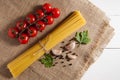 Bunch of raw spaghetti, cherry tomatoes, peppers, garlic cloves and two parsley leaves on sackcloth on a white wooden background.