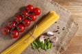 Bunch of raw spaghetti tied with rope, tomatoes cherry, slices of garlic, parsley leaves and pepper on burlap, all on brown wooden