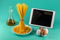 Bunch of spaghetti standing upright on a bright colored background surrounded by tablet computer, chicken eggs, olive