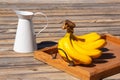 Bunch of yellow ripe bananas on a wooden tray and white jug on wooden surface on sunny day. Still life photo. Selective focus. Royalty Free Stock Photo