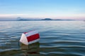 Buoy with red and white stripes on the blue water of Lake Balaton with the Badacsony mountan in the background at sunset