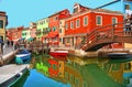 Burano island picturesque street with small colored houses, tourists on wooden bridge and beautiful water reflections