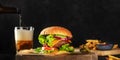 Burger and beer panorama. Hamburger with beef, cheese, onion, tomato, and green salad, a side view on a dark background Royalty Free Stock Photo