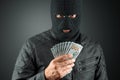 Burglar, a thug in a balaclava holds dollars in his hands on a dark background. Robbery, hacker, crime, theft. Copy space