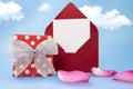 Burgundy letter with paper for text stands. Red polka dot gift box with silver bow and rose petals on blue background with clouds Royalty Free Stock Photo