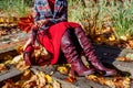 Burgundy red leather knee high boots. Fashionable woman wearing stylish blazer skirt sitting in fall park with purse Royalty Free Stock Photo