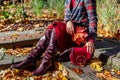 Burgundy red leather knee high boots. Fashionable woman wearing stylish blazer skirt sitting in fall park with purse Royalty Free Stock Photo