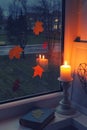 Burn candle front of window, fallen leaves and book. Moment of loneliness and autumn melancholy